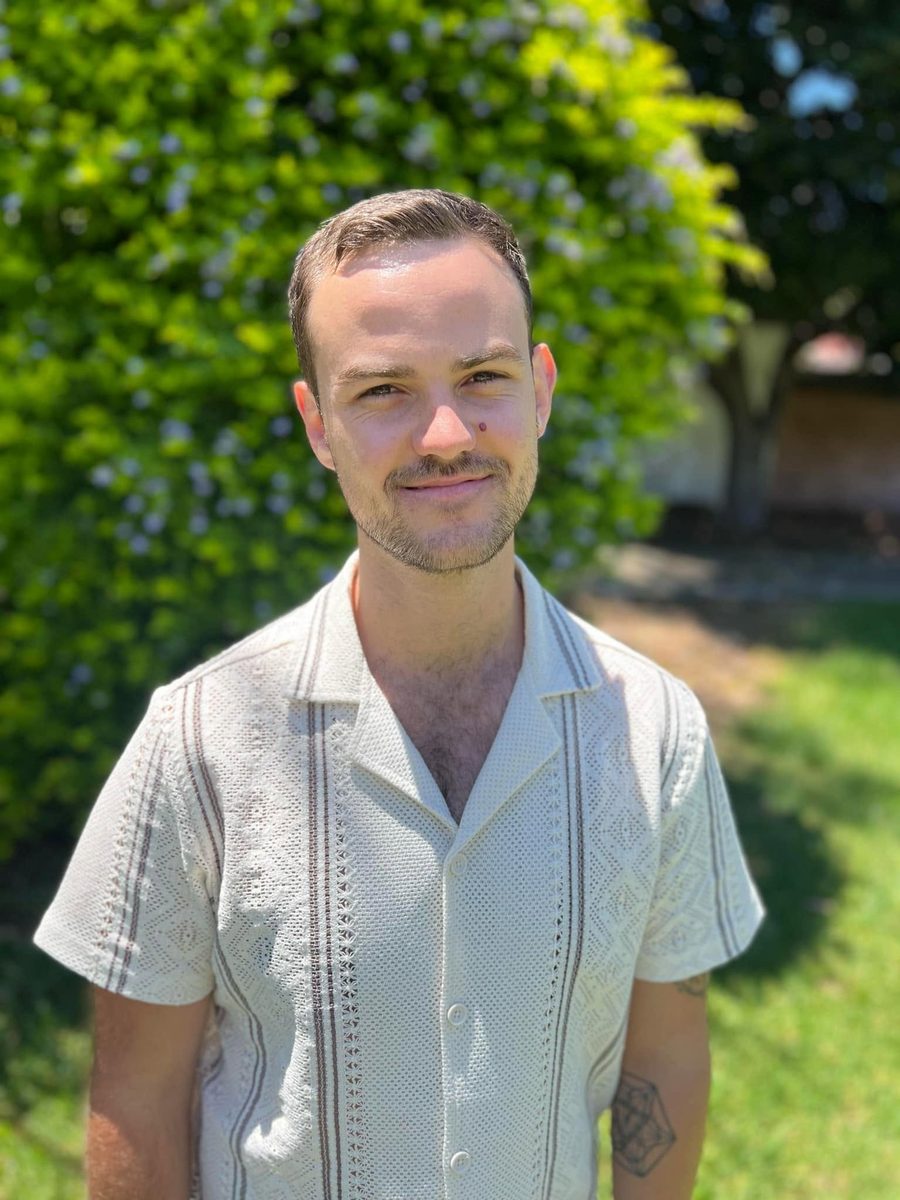 Luke Turner smiling, standing outdoors in front of a flowering hedge